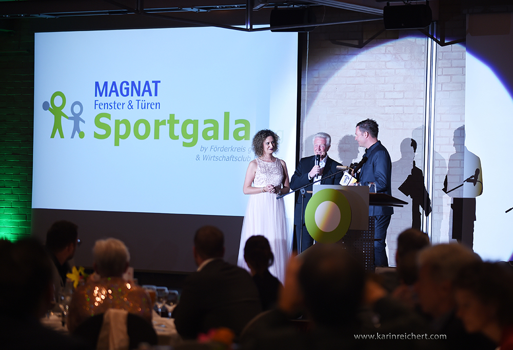 Blick aus dem Publikum auf eine Bühne, auf der drei Menschen vor dem Rednerpult stehen. Die Leinwand hinter ihnen zeigt das goolkids-Logo und die Aufschrift "Magnat-Sportgala".